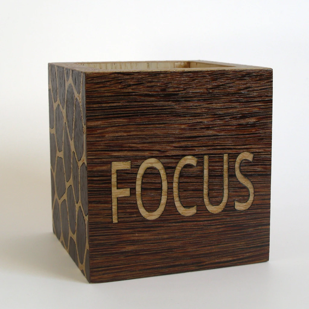 FOCUS Stiftehalter in Wenge und Eiche: dunkle Frontflaeche mit dem Schriftzug FOCUS in heller Eiche, links die Seitenflaeche mit zellartigem Muster